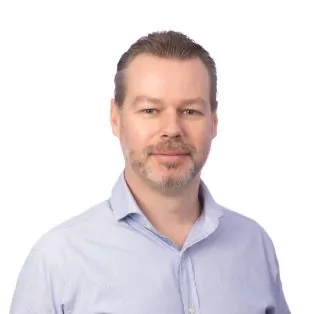 A man with light skin, short brown hair, and a trimmed beard is wearing a light blue collared shirt and posing against a plain white background. He is looking at the camera with a neutral expression.