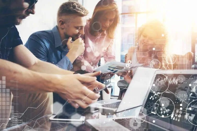 Four people collaborate around a table with laptops and documents, reviewing charts and data. Overlayed digital graphics suggest technology or data analysis in a bright, modern office setting.
